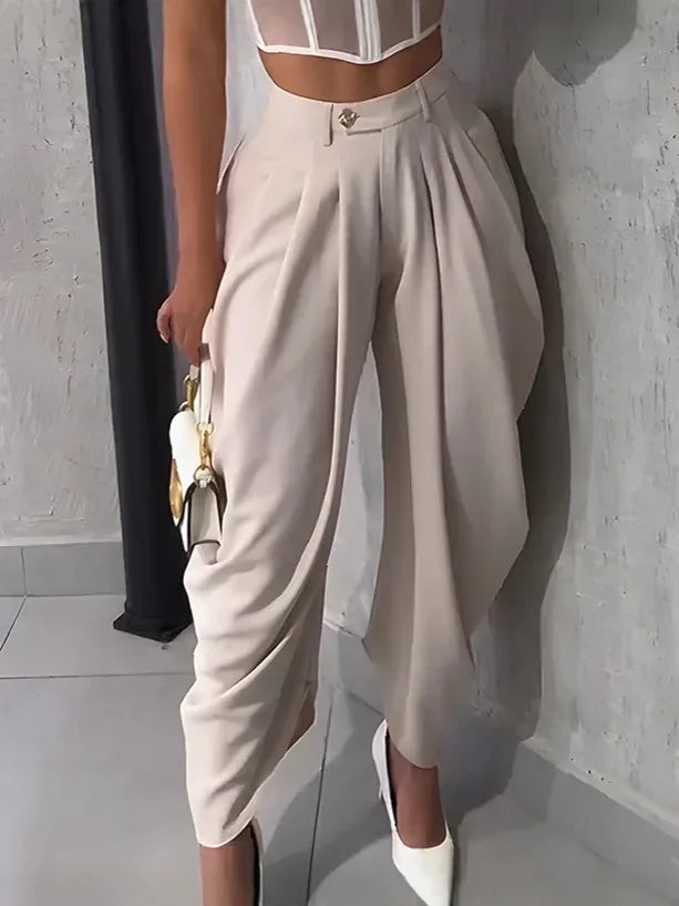 Person wearing beige high-waisted pants and a sheer top, holding a white handbag indoors.