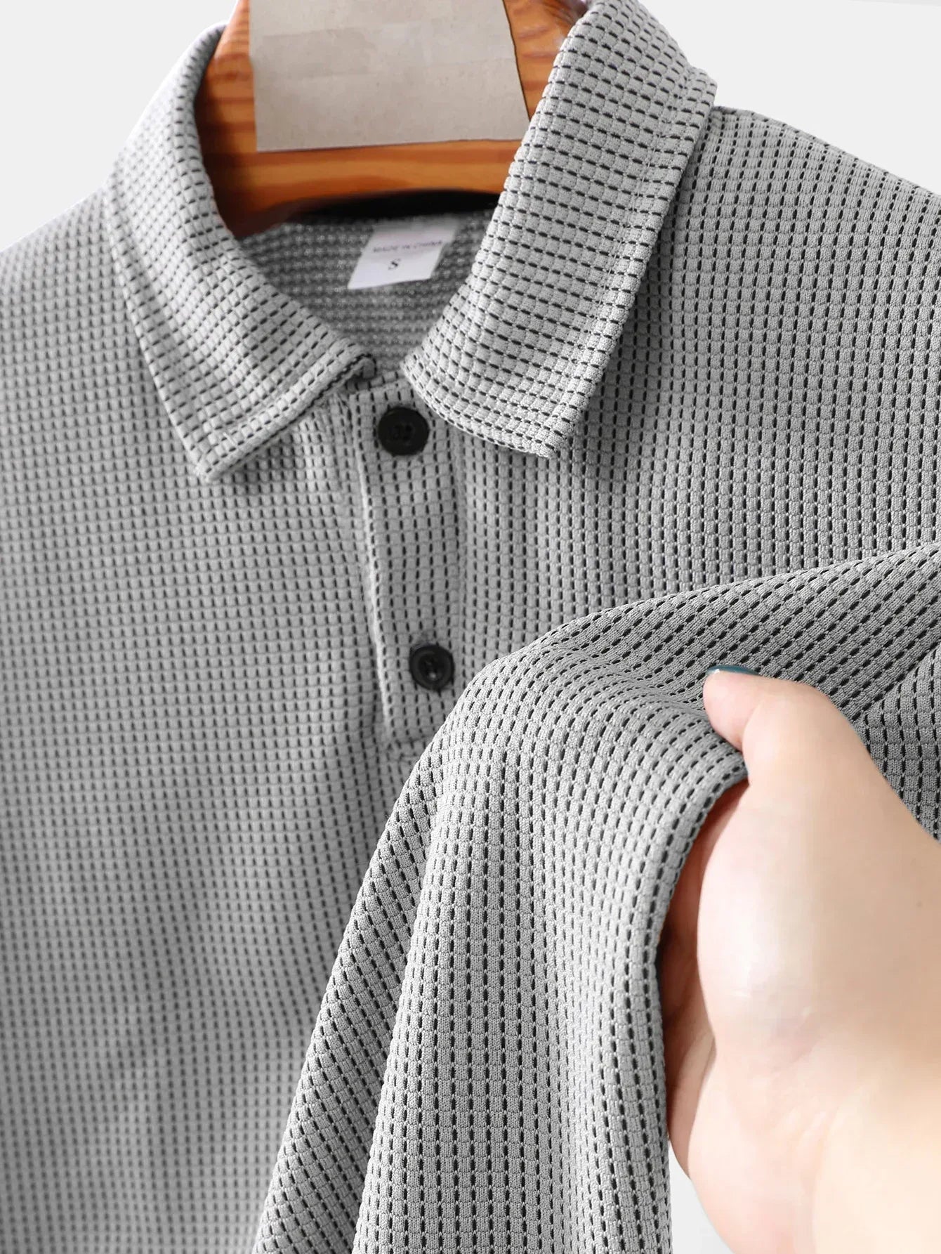 Gray textured polo shirt on a hanger with a hand touching the fabric.
