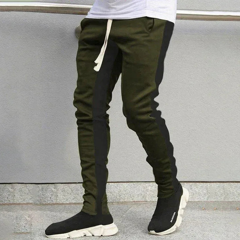 Person wearing green jogger pants with black panels and black sneakers against a gray wall.