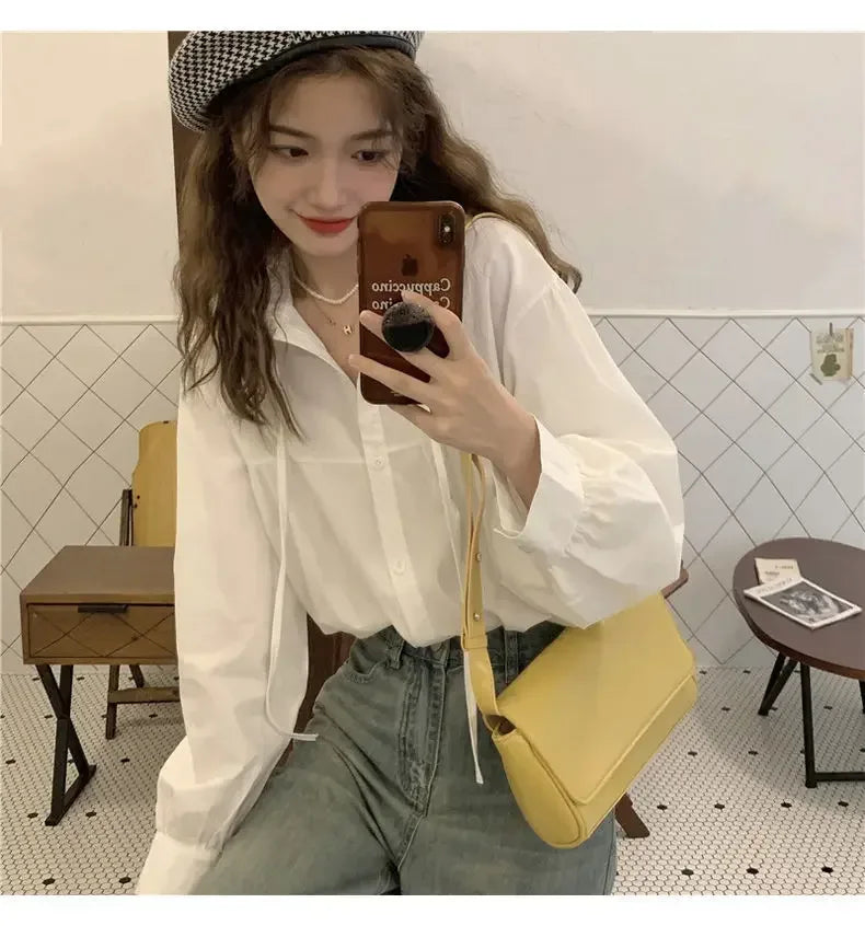 Woman taking a mirror selfie wearing a white shirt, yellow bag, and plaid hat in a tiled room.