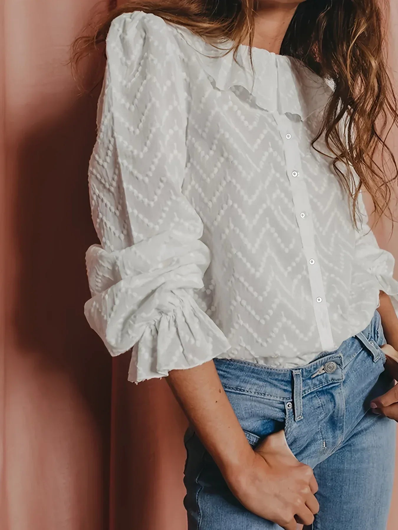 Person wearing a white patterned blouse with ruffled sleeves and blue jeans against a pink wall.